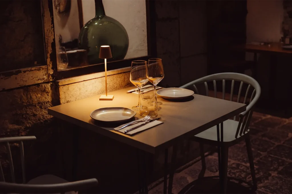 An intimate table setting for two in a dimly lit, rustic restaurant with warm table lamp lighting, wine glasses, and minimalist place settings.