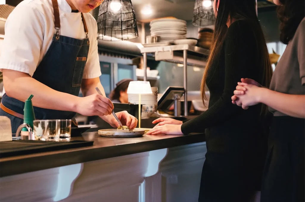 Chef using tweezers to garnish a dish for customers at a sushi restaurant counter.